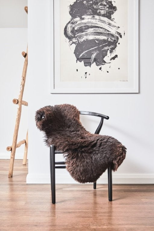 British sheepskin rug in rich dark brown draped over a black chair in a modern interior setting with timber flooring and wall art