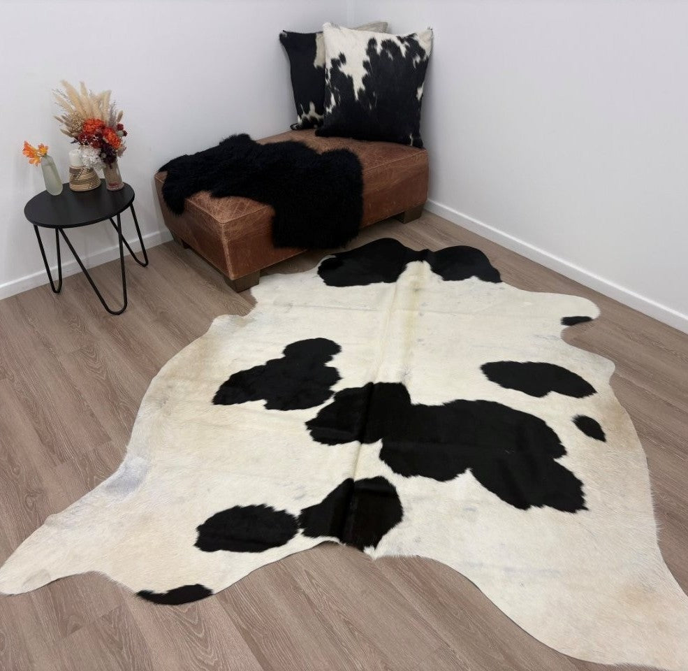 A black and white cowhide rug laid on a wooden floor beside a brown leather ottoman, a black throw blanket, a patterned cowhide cushion, and a small round side table with flowers.