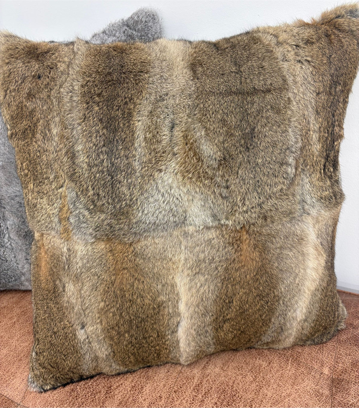 A natural brown rabbit fur cushion standing upright on a brown leather ottoman with a grey cushion behind it, positioned against a white wall.