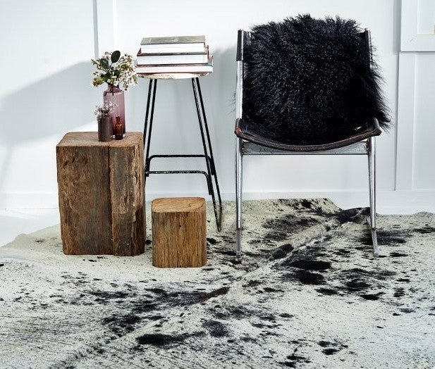 Salt and pepper cowhide rug styled in a modern interior with a chair, fur throw, wooden stools and a side table on a textured floor