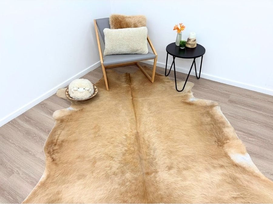 Solid beige cowhide rug styled in a living space with a chair, cushion and side table on a timber floor