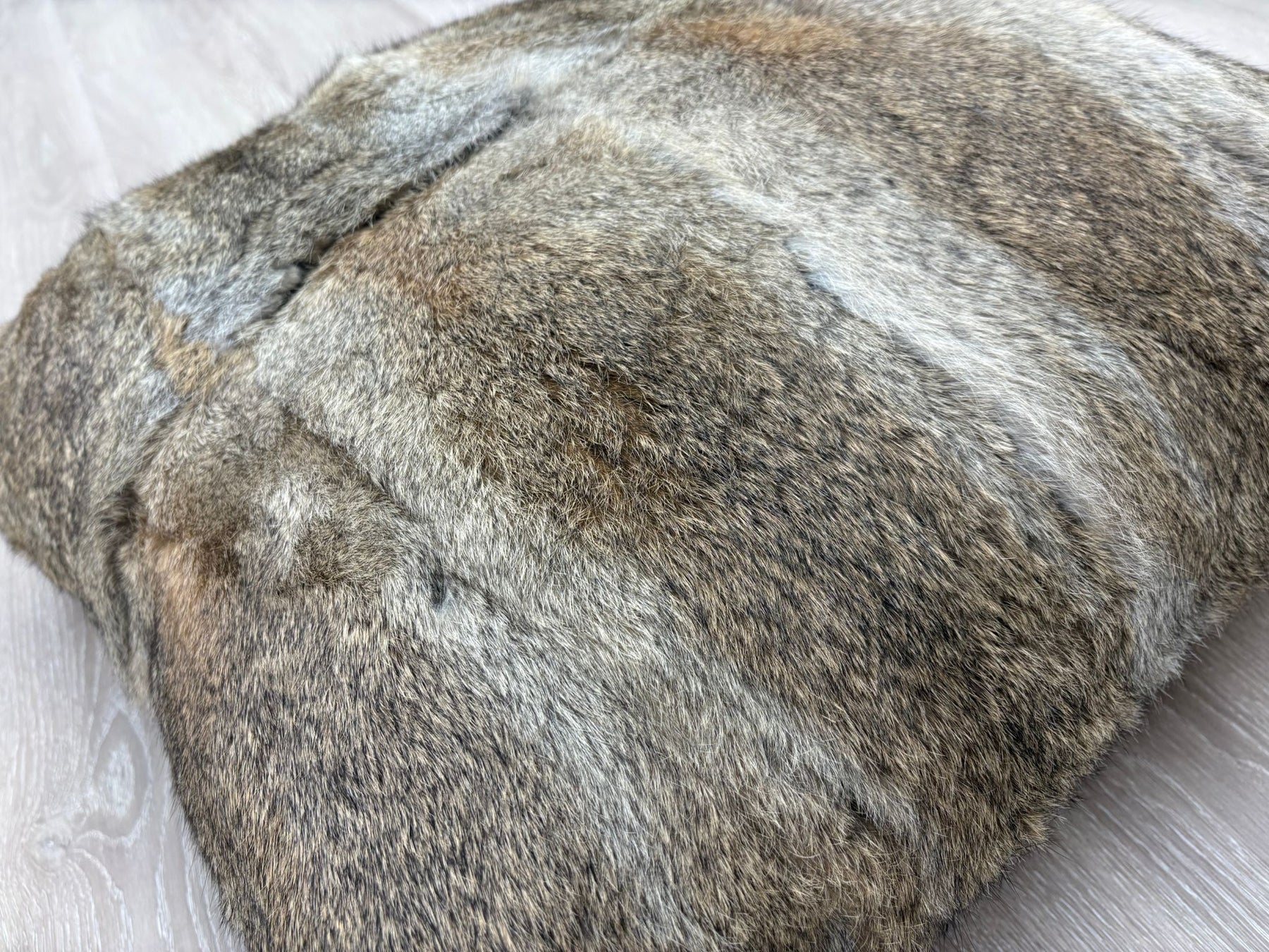 A close-up angled view of a natural brown rabbit fur cushion showing the soft fur texture and tonal variations, photographed on a light wooden floor.
