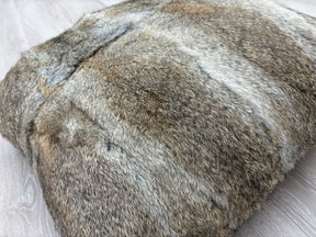 A close-up angled view of a natural brown rabbit fur cushion showing the soft fur texture and tonal variations, photographed on a light wooden floor.