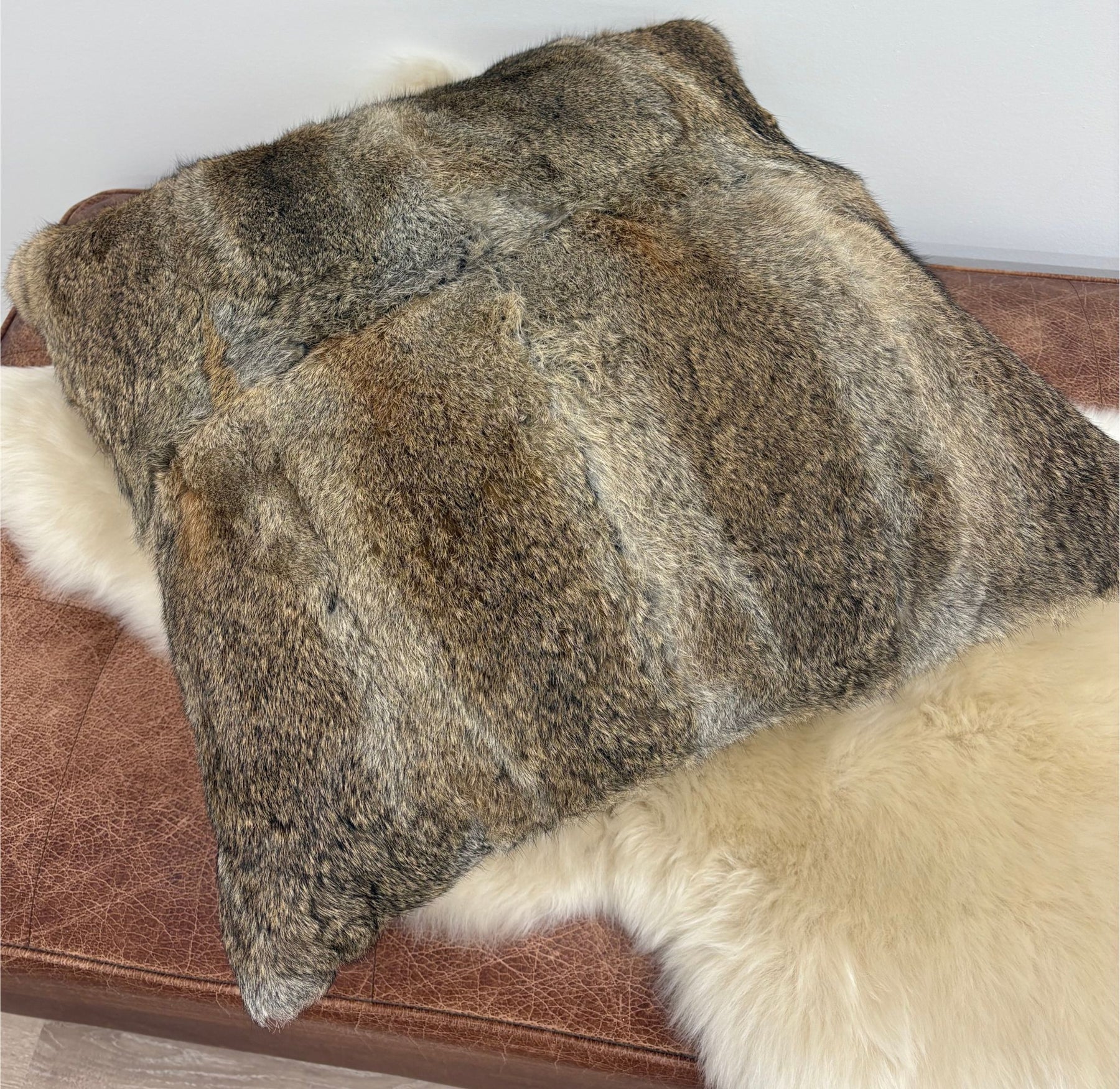 A natural brown rabbit fur cushion resting on a brown leather ottoman with a cream sheepskin rug beneath it, shown against a plain white wall.