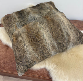 A natural brown rabbit fur cushion resting on a brown leather ottoman with a cream sheepskin rug beneath it, shown against a plain white wall.