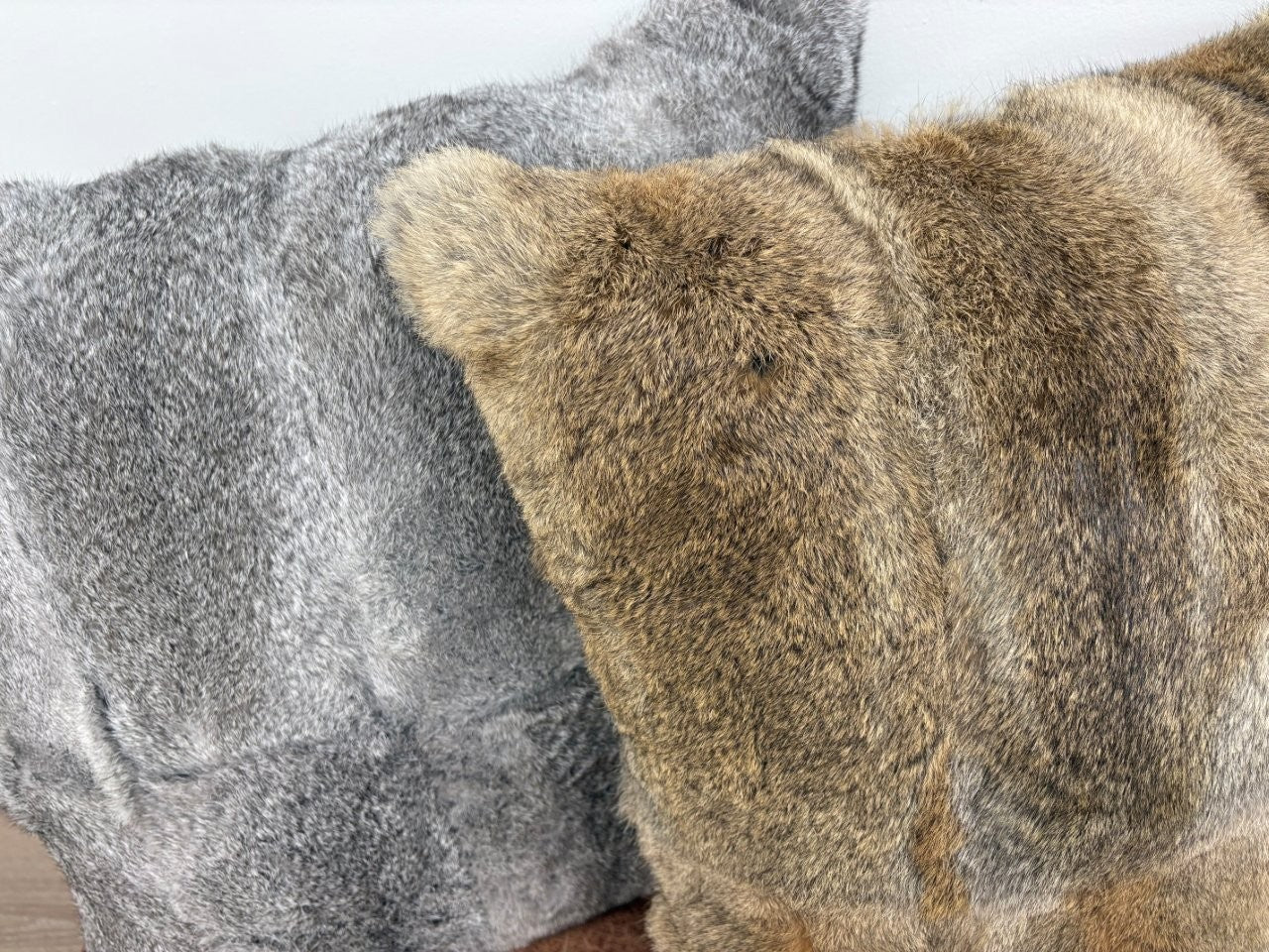 A close-up view of two rabbit fur pillows, one in grey and one in brown, placed side by side on a brown leather ottoman against a white wall.
