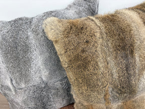 A close-up view of two rabbit fur pillows, one in grey and one in brown, placed side by side on a brown leather ottoman against a white wall.
