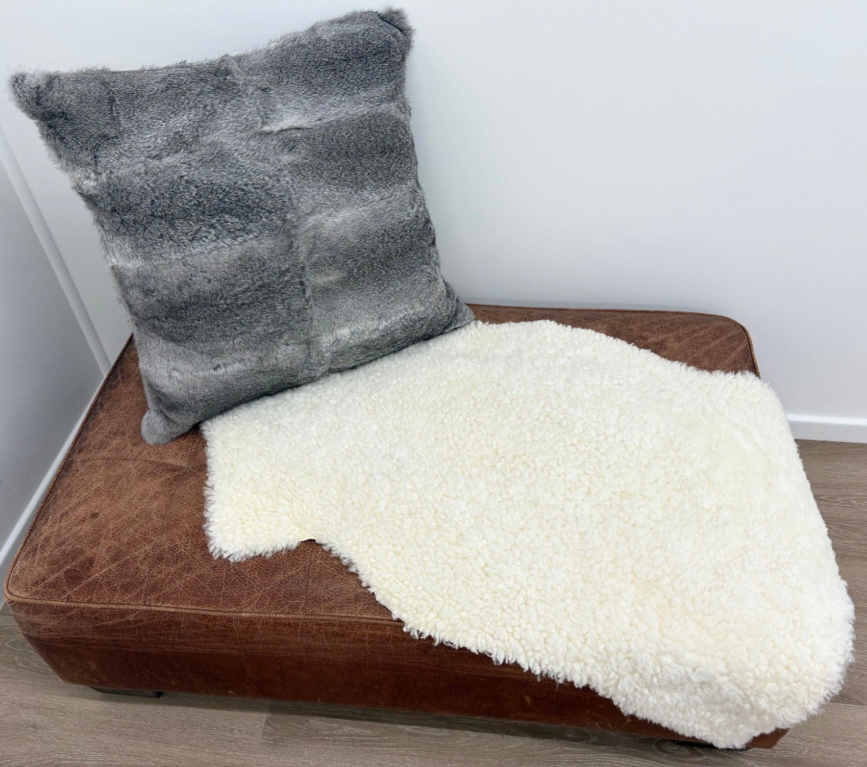 A grey rabbit fur pillow resting on a brown leather ottoman with a white sheepskin rug draped partly over the ottoman and onto a wooden floor against a white wall.