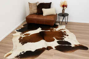 Reddish brown and white cowhide rug styled in a living room with a leather ottoman, cushions, and side table