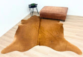 Solid brown cowhide rug styled on a light timber floor with a leather ottoman and side table