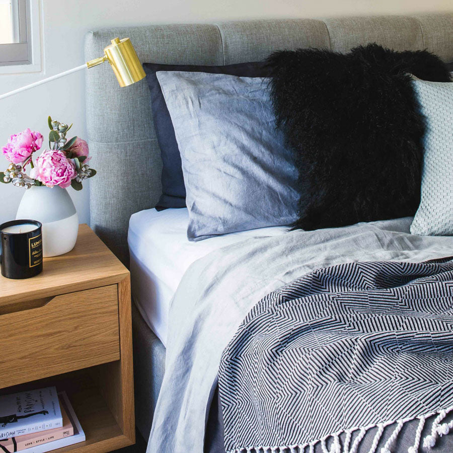 Black Mongolian sheepskin cushion styled on a bed with layered pillows and soft neutral bedding