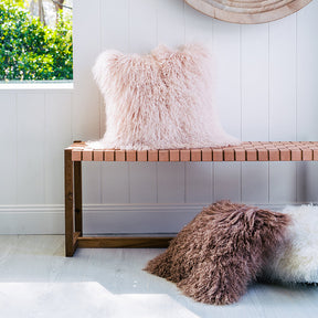 Blush Mongolian sheepskin cushion with long hair texture styled on a wooden bench in a light, neutral interior